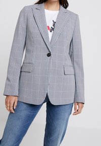 Blazer grigio e bianco a quadri con texture, colletto a notched, chiusura a un bottone e due tasche frontali, abbinato a una maglietta bianca basic.
