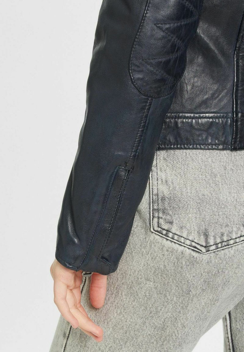 Marineblaue Lederjacke mit gesteppter Patches und Reißverschlussdetails an den Ärmeln. Über grauen Jeans getragen. Glatte Textur mit subtiler Schattierung.