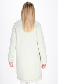 Donna con lunghi capelli biondi lisci che indossa un soprabito verde chiaro a maniche lunghe, in piedi di spalle contro uno sfondo bianco.