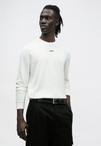 Valge pikakäistega kampsun soonikkoes mansettide ja ümara kaelusega, millel on must "HUGO" logo; kombineeritud mustade pükste ja vööga.