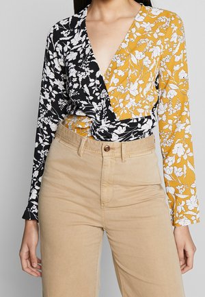Kvinde iført en snoet blomstret wrap-top med sorte og gule sektioner, kombineret med højttaljede beige bukser.