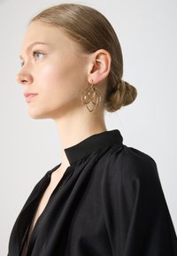 Gold hoop earrings with a layered design, featuring smooth, rounded edges. Model wears a black high-neck top. Background is plain.