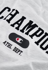 Witte t-shirt met vet zwart lettertype, "CHAMPION," met een grijze ovale logo en "ATHL. DEPT." eronder gedrukt in het zwart. Zachte stof.