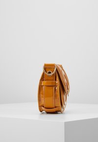 Piccola borsa a tracolla arancione lucida con motivo cucito e fibbia in metallo, mostrata in piedi su una superficie bianca contro uno sfondo semplice.