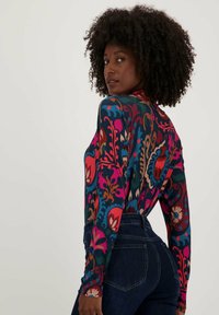 Langsluitend, langmouwshirt met een kleurrijk bloemenpatroon op een donkere achtergrond, gecombineerd met hoge donkere jeans.