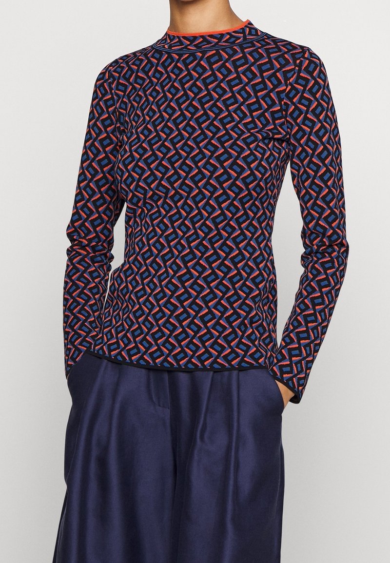 Personne portant un haut à manches longues à motifs géométriques bleu, rouge et noir avec un pantalon en satin bleu, les mains dans les poches.