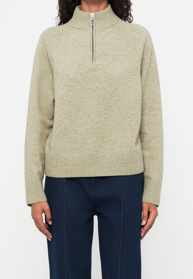 Pull beige en maille avec col montant et demi-fermeture éclair argentée à anneau, porté avec un pantalon bleu marine.