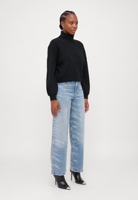 Maglione nero a collo alto, vestibilità ampia, abbinato a jeans chiari a vita alta con una texture sbiadita e una leggera brillantezza, e tacchi neri.