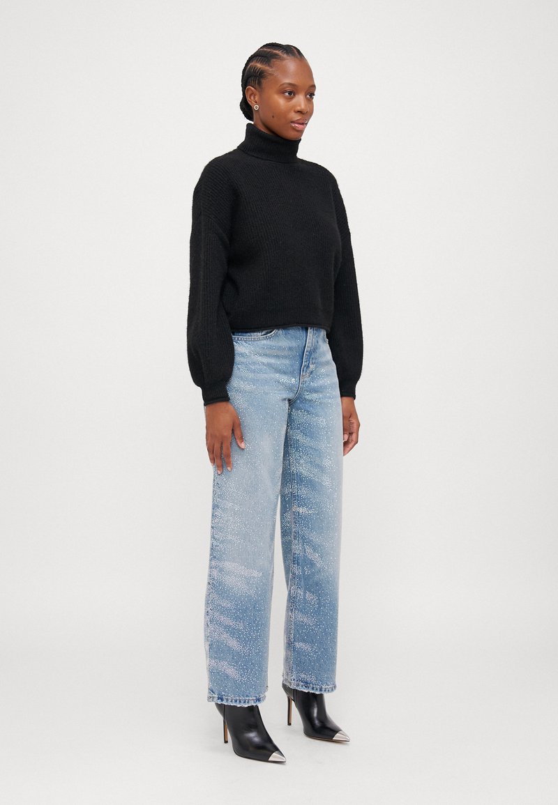 Maglione nero a collo alto, vestibilità ampia, abbinato a jeans chiari a vita alta con una texture sbiadita e una leggera brillantezza, e tacchi neri.
