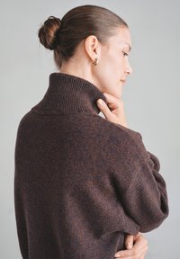 Ciemnobrązowy, teksturowany sweter z dzianiny z wysokim prążkowanym kołnierzem i opuszczonymi ramionami. Model dotyka kołnierza, z minimalną ilością dodatków.