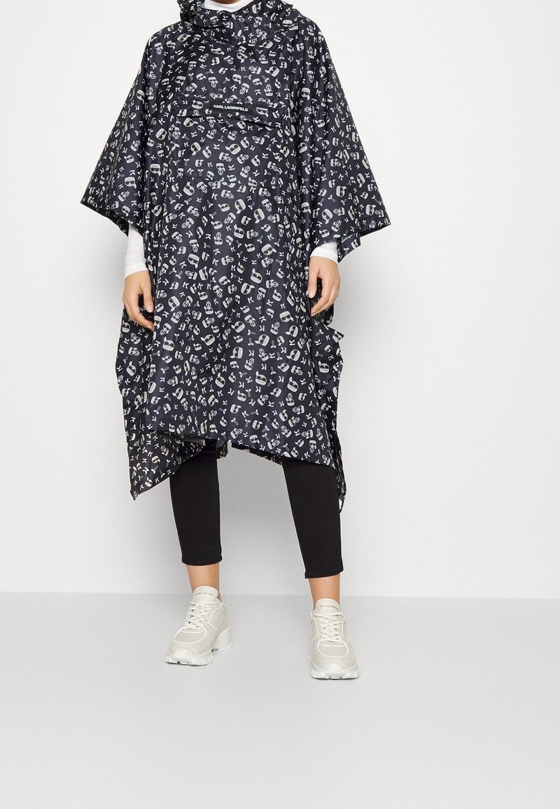 Personne portant un poncho noir et blanc à motifs avec capuche, des leggings noirs, des baskets blanches, debout devant un fond clair uni.