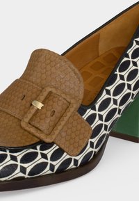 Chaussure à talon haut à motif noir et blanc avec talon bloc vert, bride à boucle beige texturée et semelle intérieure marron rembourrée.