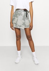 Falda de camuflaje en tonos verdes claros, con un bolsillo lateral, trabillas para el cinturón y un dobladillo recto, combinada con zapatillas blancas y calcetines.