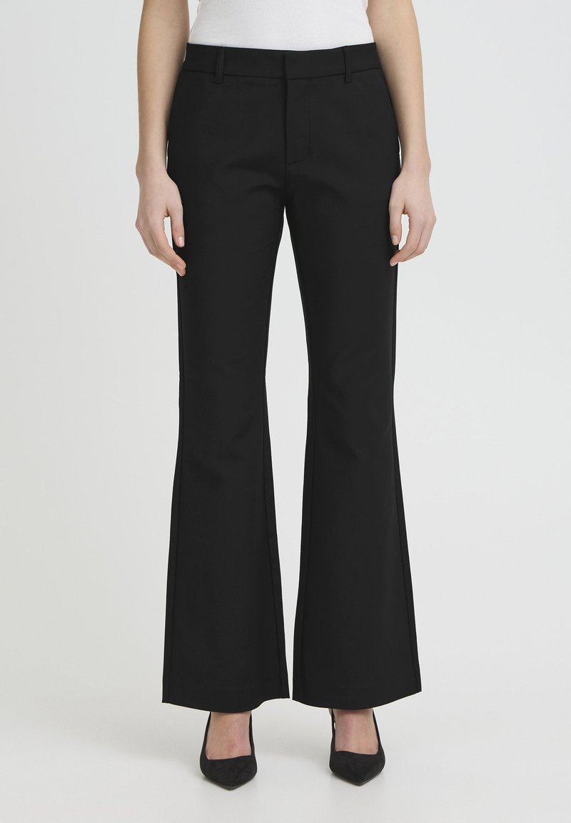 Femme portant un pantalon évasé taille haute noir avec poches latérales et des escarpins noirs à bout pointu, debout devant un fond clair uni.