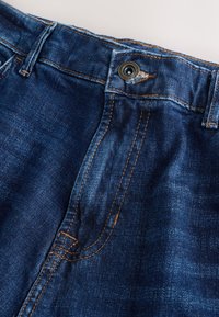 Gros plan d’un jean en denim bleu foncé montrant la ceinture, le bouton, les passants de ceinture et les détails de couture orange.