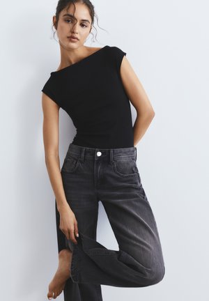 Femeie îmbrăcată într-un top negru fără mâneci, mulat, și blugi negri estompați, ținând un picior de pantaloni în timp ce se sprijină ușor de un perete alb.