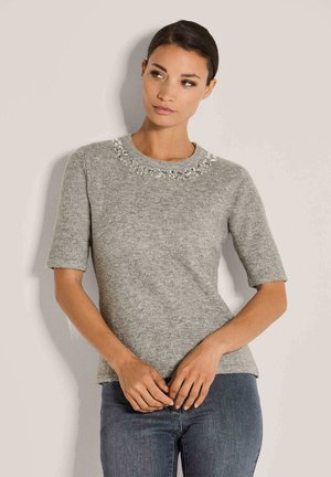 Eine Frau trägt einen grauen Kurzarm-Pullover mit einem mit Juwelen besetzten Ausschnitt und kombiniert ihn mit dunkelblaue Jeans. Sie steht vor einer hellen Wand.