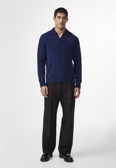 Mies, joka seisoo mustissa housuissa, mustissa kengissä ja pitkähihnaisessa tummansinisessä kauluspaidassa valkoista taustaa vasten.