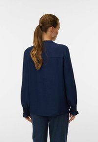 Donna con lunga coda di cavallo, che indossa una camicia blu navy a maniche lunghe e pantaloni scuri, girata di spalle su uno sfondo bianco semplice.