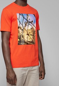 Pomarańczowy bawełniany T-shirt z dużym fotograficznym nadrukiem przedstawiającym pustynną scenerię z palmami, oprawiony czarnym wzorem przypominającym ogniwa łańcucha.