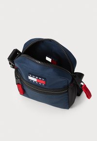 Borsa di stoffa blu navy con chiusura a zip, dettagli rossi e logo a patch sul davanti. Interno foderato con un materiale liscio; design compatto e versatile.