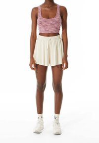 Personne portant un haut court sans manches rose, un short ample crème avec cordon de serrage, des chaussettes blanches et des baskets blanches, debout devant un fond blanc.