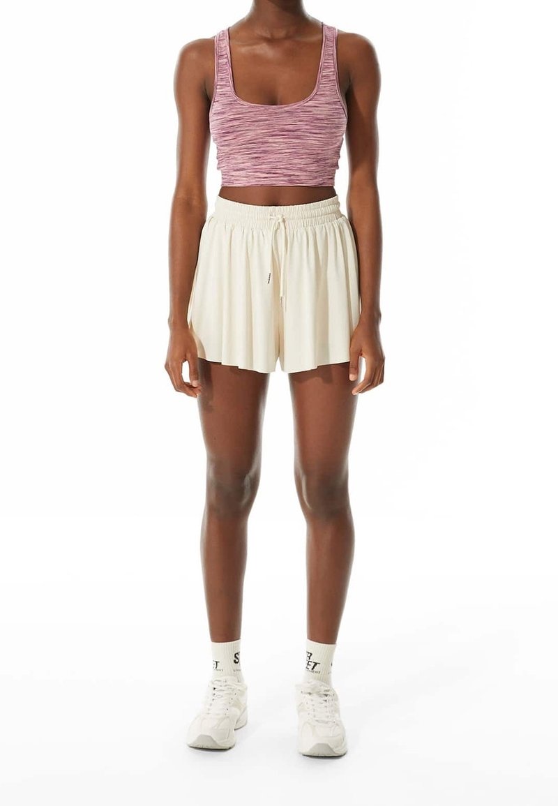 Personne portant un haut court sans manches rose, un short ample crème avec cordon de serrage, des chaussettes blanches et des baskets blanches, debout devant un fond blanc.