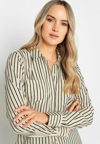 Camisa beige de botones con rayas verticales negras. Presenta un cuello clásico, un bolsillo en el pecho izquierdo y mangas largas con puños abotonados.