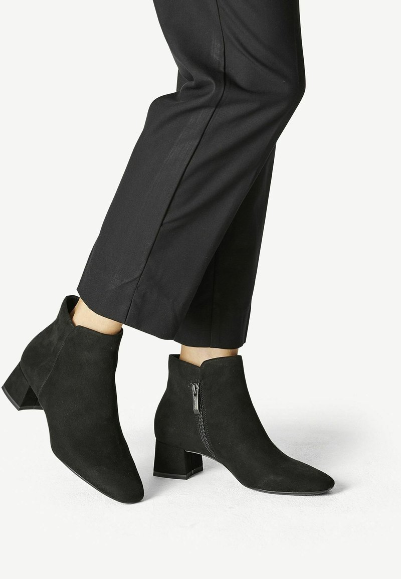 Personne portant des bottines noires à talons carrés et fermetures éclair latérales, associées à un pantalon noir court sur un fond uni.