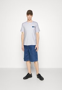 Svijetlosivi t-shirt kratkih rukava s plavim i crnim logotipom, uparen s plavim trapericama do koljena i crnim tenisicama, te smešnim kapom.