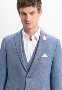 Lichtblauwe textuur blazer met twee knopen, wit overhemd en witte zakdoek in de pocket. Decoratieve bloem pin op de revers.