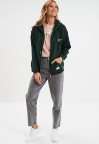 Grüne Zip-Up-Hoodie mit goldenem Logo, getragen über einem hellrosa Shirt, kombiniert mit grauen Jeans und weißen Turnschuhen. Lässig, aus weichem Stoff.