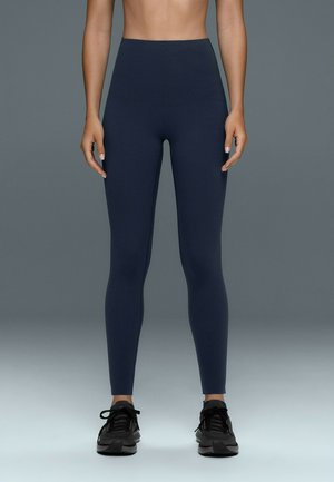 Personne portant des leggings bleu foncé taille haute et des chaussures de sport noires, debout devant un fond gris uni.