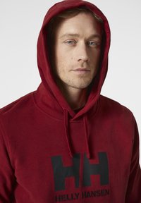 Červená bavlněná mikina s kapucí a přední kapsou, s velkým černým logem Helly Hansen. Má stahovací kapuci a jemnou texturu.