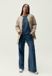 Prošívaná béžová bomberová bunda přes modrou mikinu a odpovídající rozšířené kalhoty. Tento outfit se vyznačuje hladkými texturami a sportovním designem.