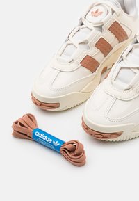 Valkoiset urheilukengät, joissa on teksturoitu beigen värinen koriste ja kumipohjat. Mukana on kanelinvärinen nauha, joka on pakattu sinisellä brändäyksellä.