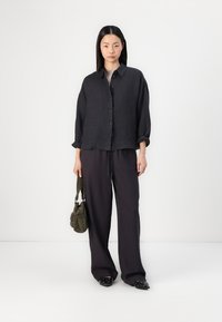 Black linen button-up shirt with rolled sleeves, paired with wide-leg dark trousers. Model carries a small green handbag, wearing black flats.