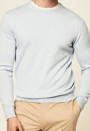 Mężczyzna ubrany w jasnoniebieski sweter z okrągłym dekoltem na białej koszulce pod spodem oraz beżowe spodnie, z jedną ręką w kieszeni.