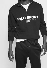 Schwarze Zip-Up-Sweatjacke mit "POLO SPORT"-Aufdruck und Logo in Weiß, kombiniert mit schwarzen Jogginghosen mit Kordelzug. Weiches Material, lockere Passform.