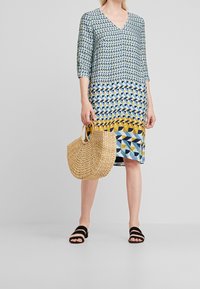 Vestido estampado con escote en V y mangas de tres cuartos, que presenta un diseño geométrico en azul, amarillo y blanco. Sostiene un bolso de paja trenzada.