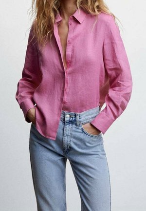 Mujer con una camisa holgada rosa abotonada, con las mangas bajadas, y jeans de tiro alto azul claro, con las manos en los bolsillos.