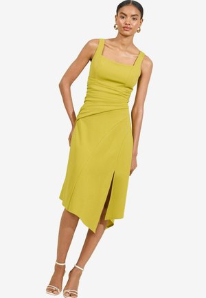 Yellow sleeveless dress with a square neckline, ruched texture, asymmetrical hem, and side slit. Paired with clear strap heels.