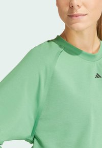 Zelené sportovní tričko s kulatým výstřihem, volným střihem a krátkými rukávy. Má černé logo akcenty poblíž výstřihu. Hladká, měkká textura.