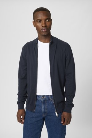 Navy knit cardigan with a stand collar and zipper closure, featuring ribbed cuffs and hem. Worn over a white t-shirt and denim jeans.