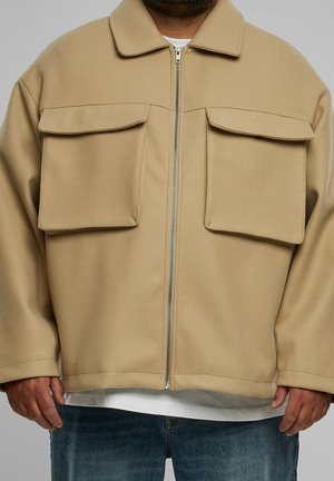 Mann trägt eine beige Reißverschlussjacke mit großen Klappentaschen über einem weißen Hemd und blauen Jeans, Hände entspannt an den Seiten.