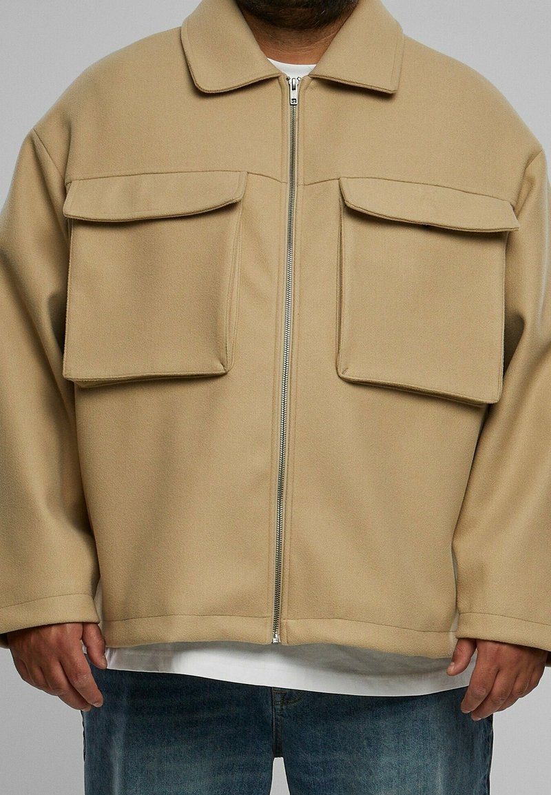 Homme portant une veste beige zippée avec de grandes poches à rabat sur une chemise blanche et un jean bleu, mains détendues le long du corps.