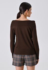 Brauner off-shoulder Strickpullover mit langen Ärmeln, geripptem Halsausschnitt, kombiniert mit grauen karierter Shorts. Glatte Textur, figurbetontes Design.