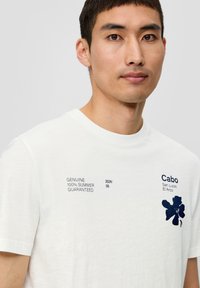 Biała koszulka z krótkim rękawem z niebieskim wzorem kwiatowym i elementami tekstowymi. Miękki materiał z luźnym krojem. Prosty i estetyczny design.