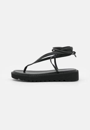 Sort flad sandal med tyk sål og flere tynde ankelremme bundet i knude, set fra siden på en ensfarvet hvid baggrund.