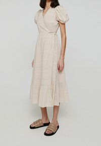 Robe portefeuille beige clair avec manches bouffantes, longueur midi, ourlet à volants et tissu texturé. Associée à des sandales camel à enfiler.